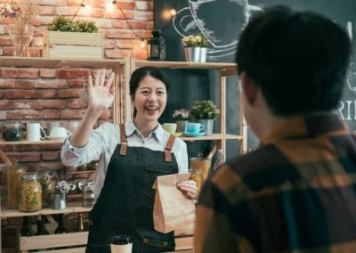 A smiling cafe owner greets a customer and hands them a packed food. A F&B POS system is on the counter, helping to streamline the cafe's operation.