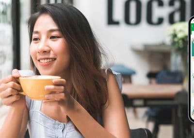 Customer Loyalty & EQuip Rewards: Smiling woman sipping coffee at a cafe with a mock-up phone displaying Edgeworks' customer loyalty & reward program.