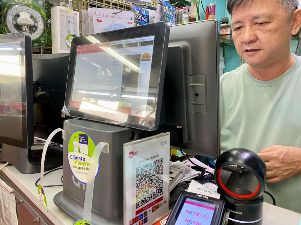 A point-of-sale (POS) system setup at TS Yong Trading, showing multiple touchscreen displays and payment terminals. A Climate Voucher acceptance sticker is prominently displayed on the POS terminal, and a QR code for payments is visible. A NETS payment device sits at the counter alongside a Honeywell barcode scanner. 