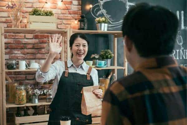 A smiling cafe owner greets a customer and hands them a packed food. A POS system is on the counter, helping to streamline the cafe's operation.