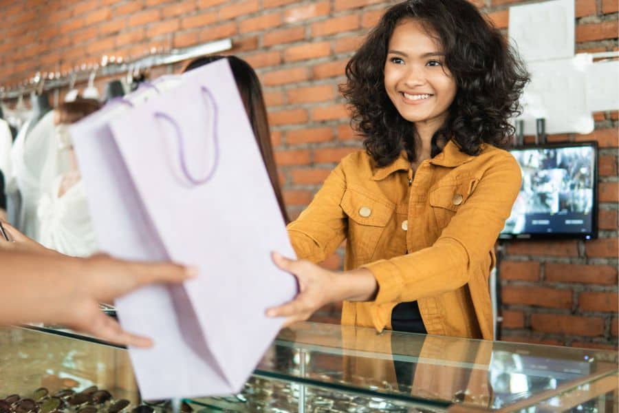 A smiling salesgirl at the counter handing over purchased goods to a customer, showcasing the enhanced customer experience with a retail POS system.