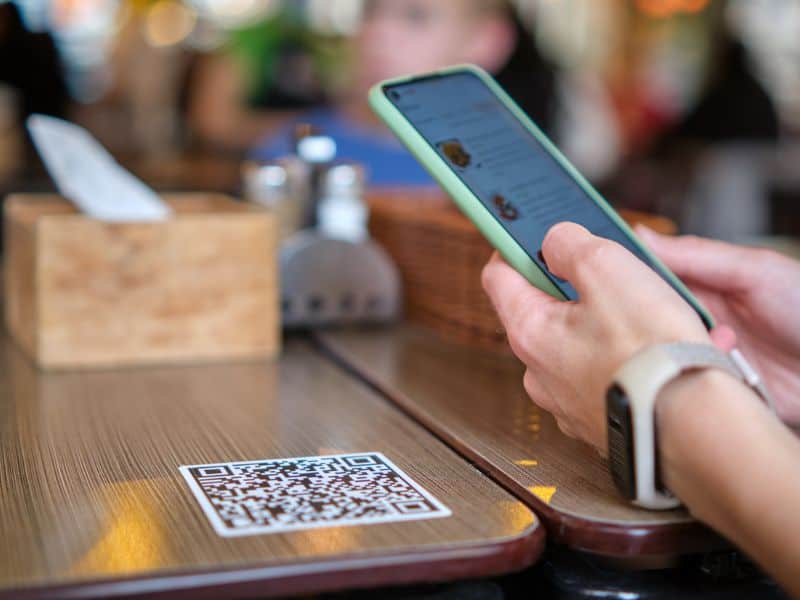 A woman sitting at a restaurant table using her smartphone to scan a QR code on the table to access the restaurant's digital menu. This is possible thanks to a restaurant POS system that allows contactless menus and payments.